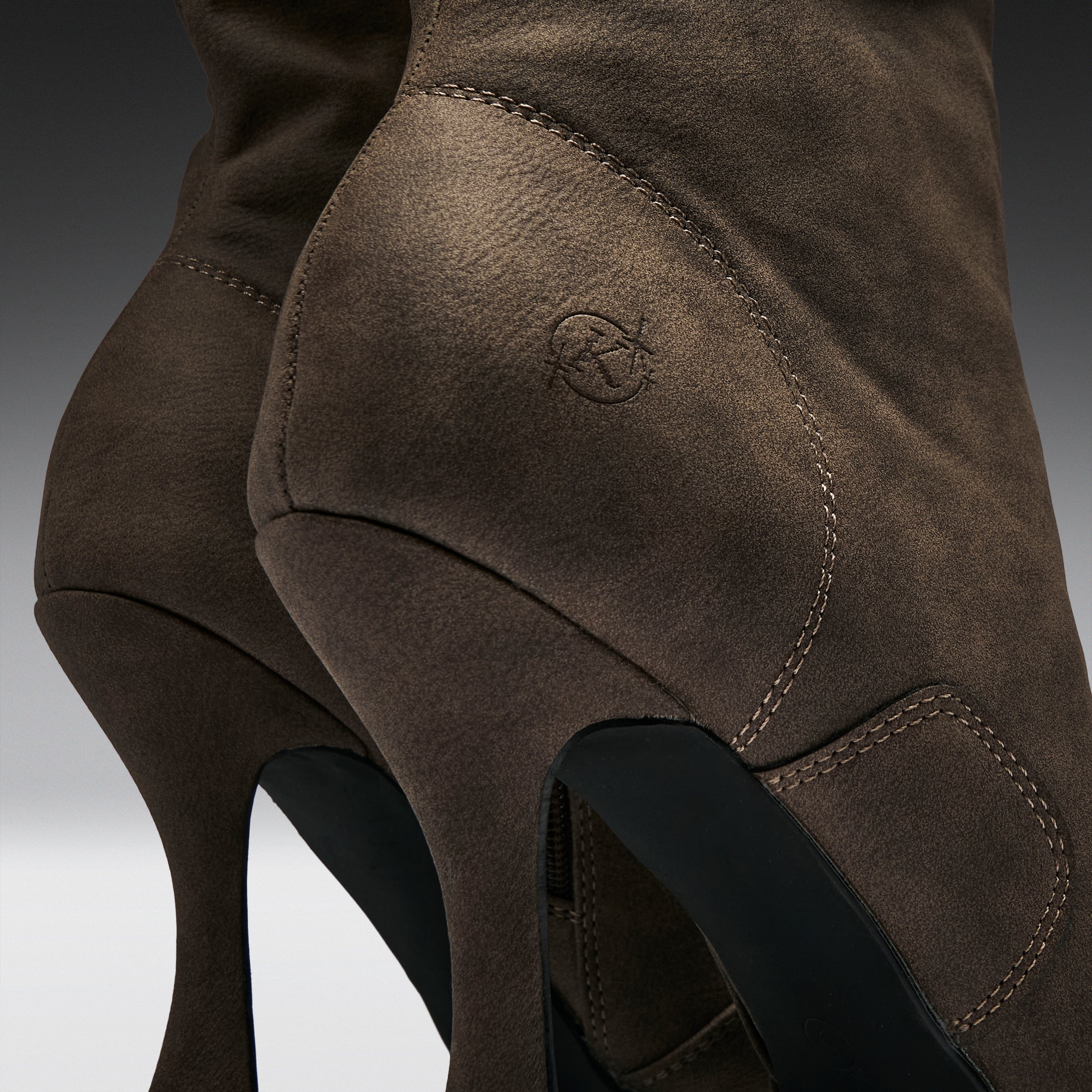 Close-up of a brown suede high-heeled boot showing the side heel, stitching, and a small embossed logo on a dark gray gradient background.