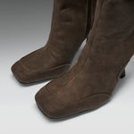 A close-up of a pair of brown suede over-the-knee boots with a square toe and low block heel, shown on a neutral gray background.