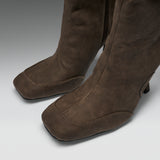 A close-up of a pair of brown suede over-the-knee boots with a square toe and low block heel, shown on a neutral gray background.
