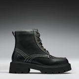 A single black leather lace-up boot with contrast white stitching, gold-tone eyelets, and a chunky lug sole, photographed on a dark-to-light gray gradient studio background.