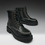 A pair of black leather lace-up boots with chunky lug soles, contrast white stitching, and brass eyelets, photographed on a dark-to-light gray gradient studio background.