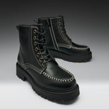 A pair of black leather lace-up boots with chunky lug soles, contrast white stitching, and brass eyelets, photographed on a dark-to-light gray gradient studio background.