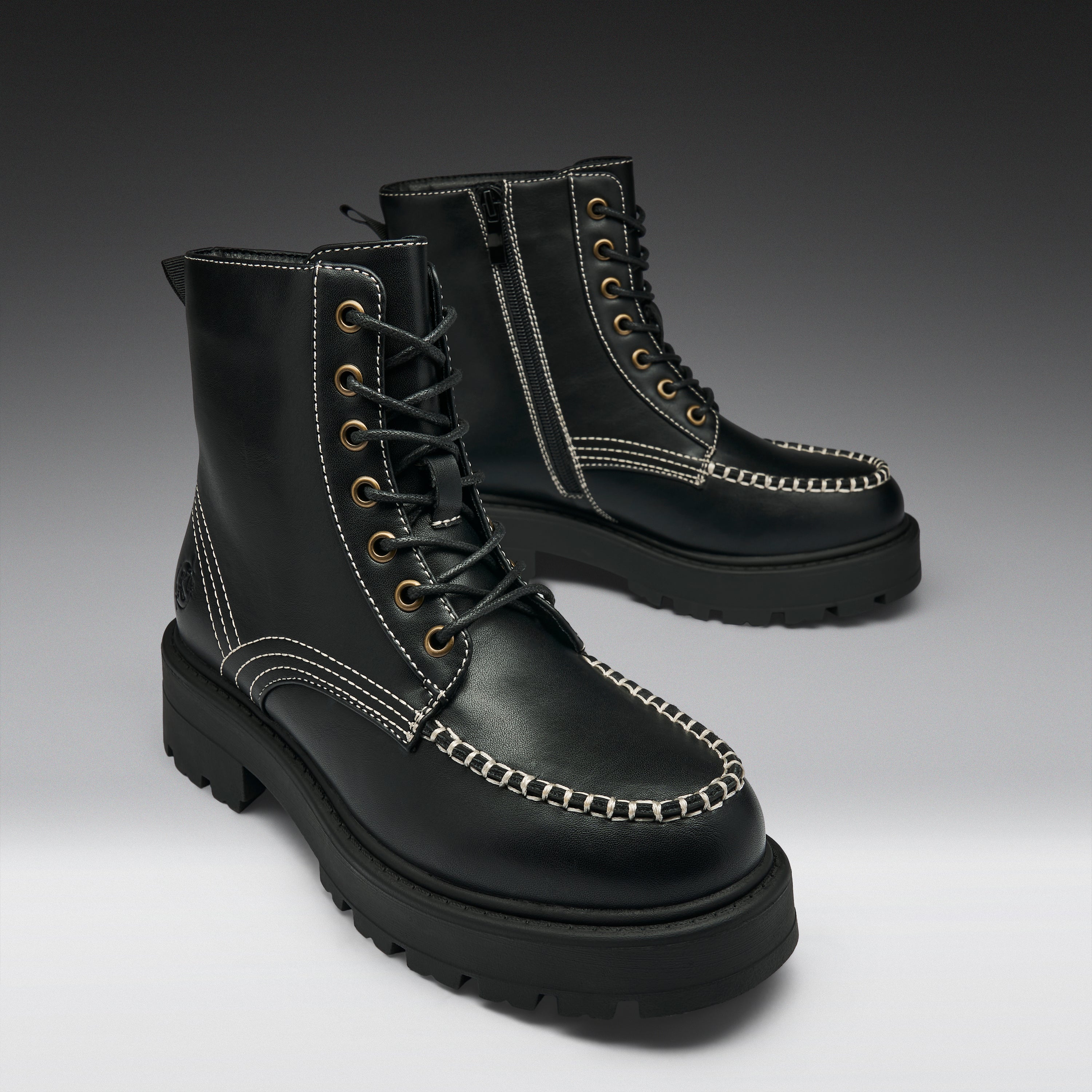 A pair of black leather lace-up boots with chunky lug soles, contrast white stitching, and brass eyelets, photographed on a dark-to-light gray gradient studio background.