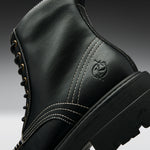 Close-up of a black leather lace-up boot with white contrast stitching, gold eyelets, and an embossed circular logo on the side, shown against a dark gray gradient background.