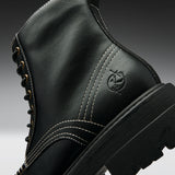 Close-up of a black leather lace-up boot with white contrast stitching, gold eyelets, and an embossed circular logo on the side, shown against a dark gray gradient background.