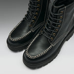 A pair of black leather lace-up boots with distinctive contrast white stitching and silver chain trim along the toe, shown on a light gray gradient background.