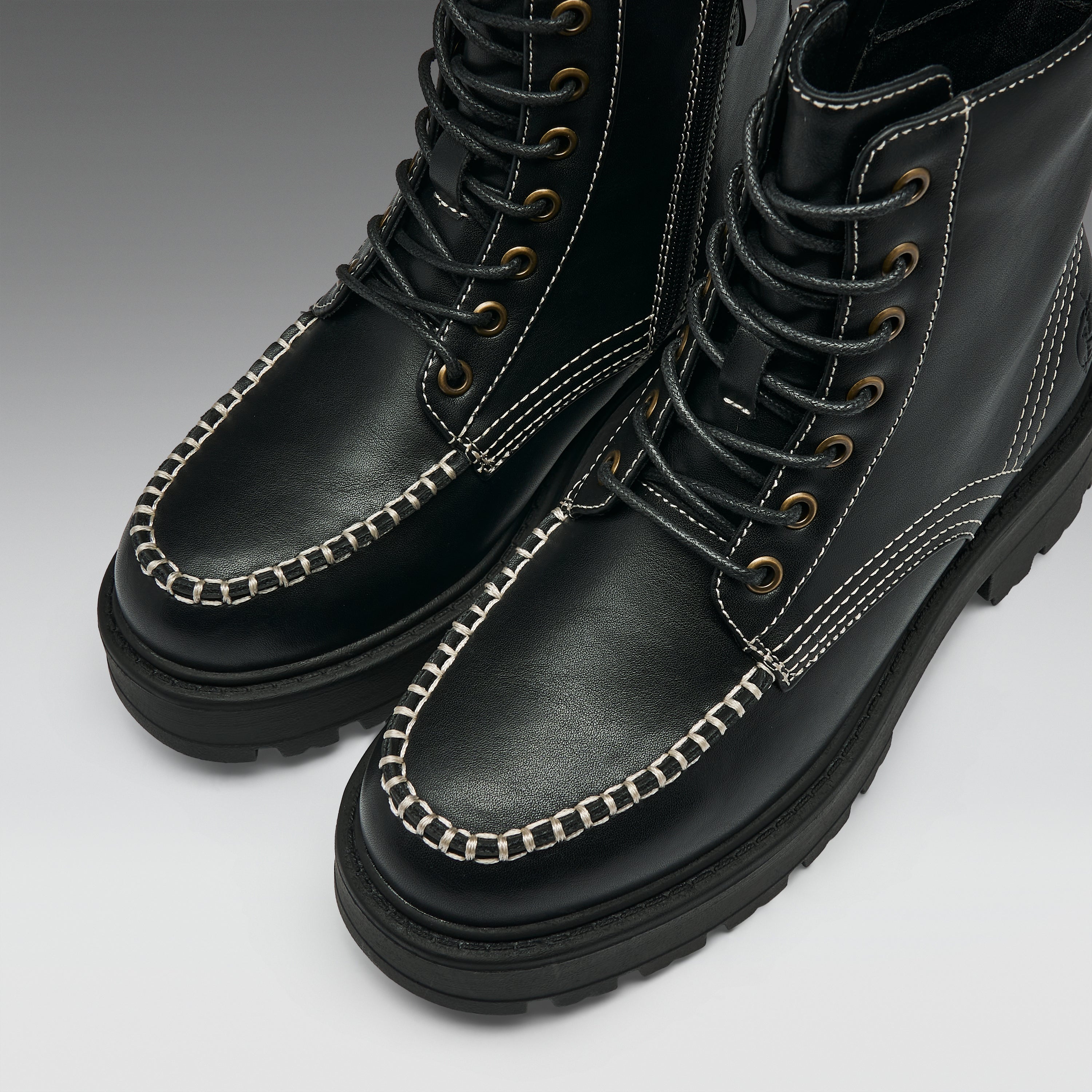 A pair of black leather lace-up boots with distinctive contrast white stitching and silver chain trim along the toe, shown on a light gray gradient background.