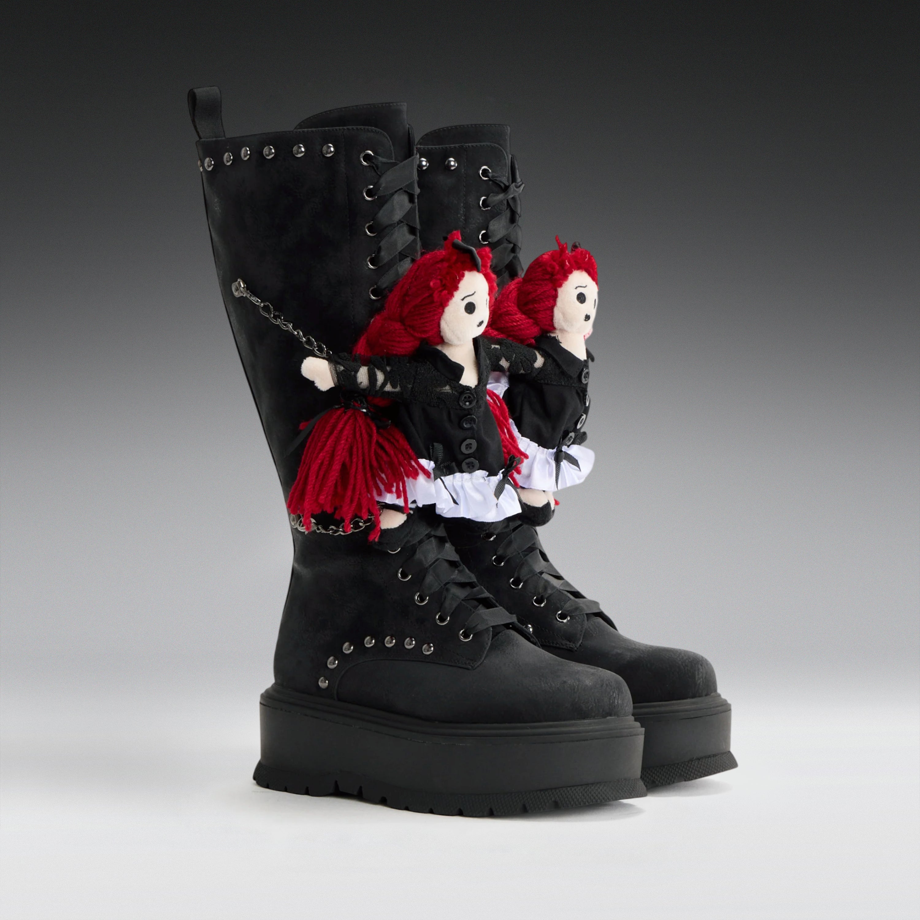 A pair of black lace-up combat boots with chunky platform soles, decorated with two red-haired doll figures dressed in black and white outfits attached near the upper shafts, photographed on a smooth gray studio background.