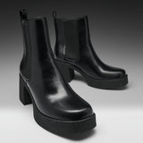 A pair of black leather Chelsea-style ankle boots with platform soles and block heels, photographed on a clean gray studio background.