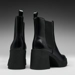 A pair of black leather Chelsea-style ankle boots with chunky block heels, shown from the back and side against a dark gray gradient studio background.