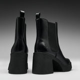 A pair of black leather Chelsea-style ankle boots with chunky block heels, shown from the back and side against a dark gray gradient studio background.
