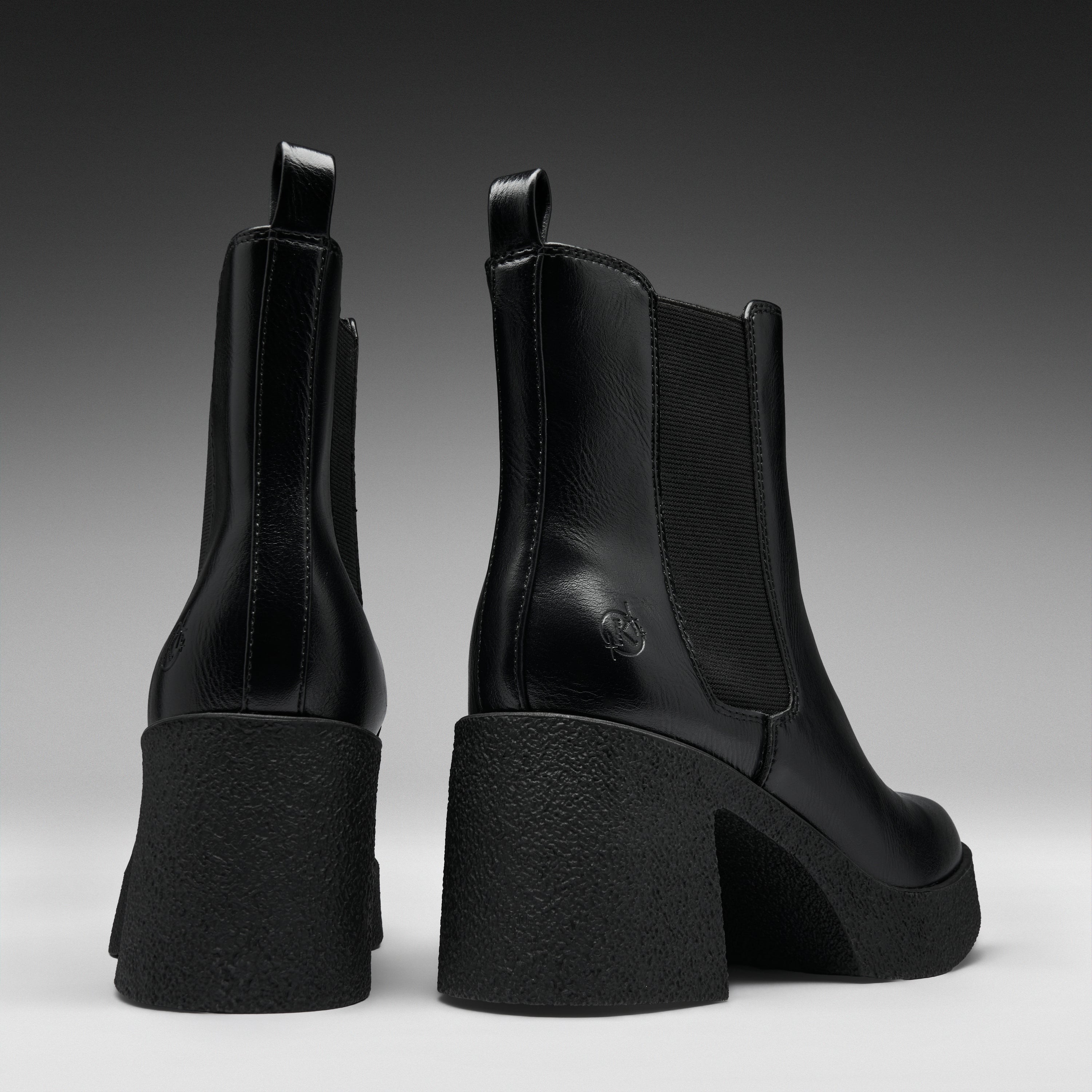 A pair of black leather Chelsea-style ankle boots with chunky block heels, shown from the back and side against a dark gray gradient studio background.