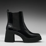 A black leather ankle boot with a chunky block heel and platform sole, featuring a black elastic side panel and a small embossed logo on the side.