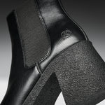 Close-up of a black leather ankle boot with a chunky textured block heel and a tonal embossed logo on the side.