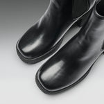 A close-up studio shot of a pair of black leather ankle boots with square toes and elastic side panels, photographed on a light gray gradient background.