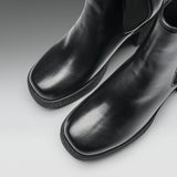 A close-up studio shot of a pair of black leather ankle boots with square toes and elastic side panels, photographed on a light gray gradient background.