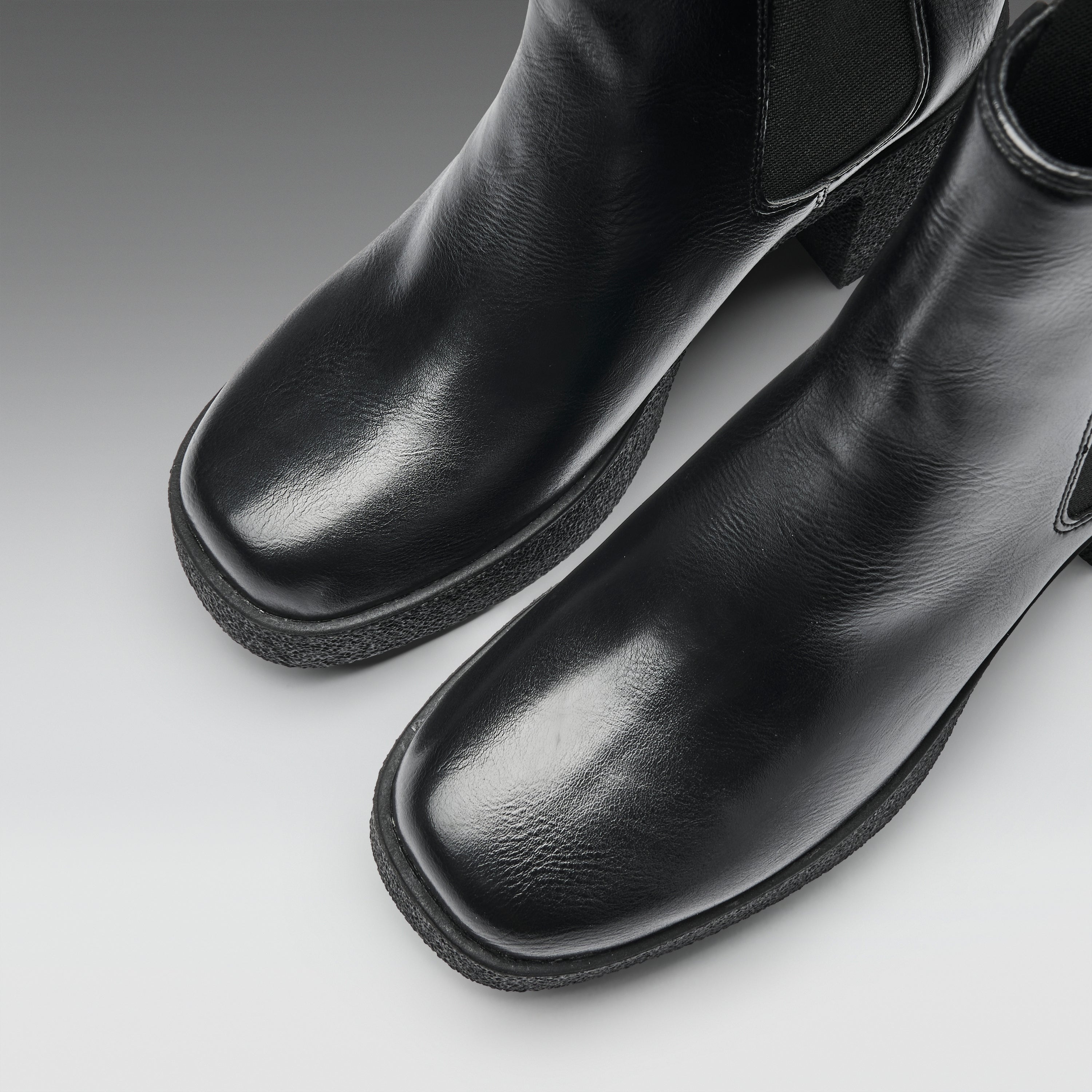 A close-up studio shot of a pair of black leather ankle boots with square toes and elastic side panels, photographed on a light gray gradient background.