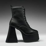 A single black leather lace-up ankle boot with a dramatic high block heel and platform sole, shown in profile against a dark gray gradient studio background.