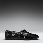 A black leather loafer flat decorated with silver-tone metal eyelets and dangling charms shaped like a star, bow, key, and heart, shown against a dark-to-light gray gradient studio background.