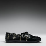 A black leather loafer flat decorated with silver-tone metal eyelets and dangling charms shaped like a star, bow, key, and heart, shown against a dark-to-light gray gradient studio background.
