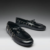 A pair of glossy black leather loafers decorated with silver-tone metal eyelets, tassels, and hanging charms, displayed on a dark-to-light gray gradient studio background.