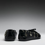 A pair of black patent leather lace-up flats decorated with silver-tone metal charms, including a star, cross, and clover, shown from the back and side against a dark gray gradient background.