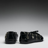 A pair of black patent leather lace-up flats decorated with silver-tone metal charms, including a star, cross, and clover, shown from the back and side against a dark gray gradient background.