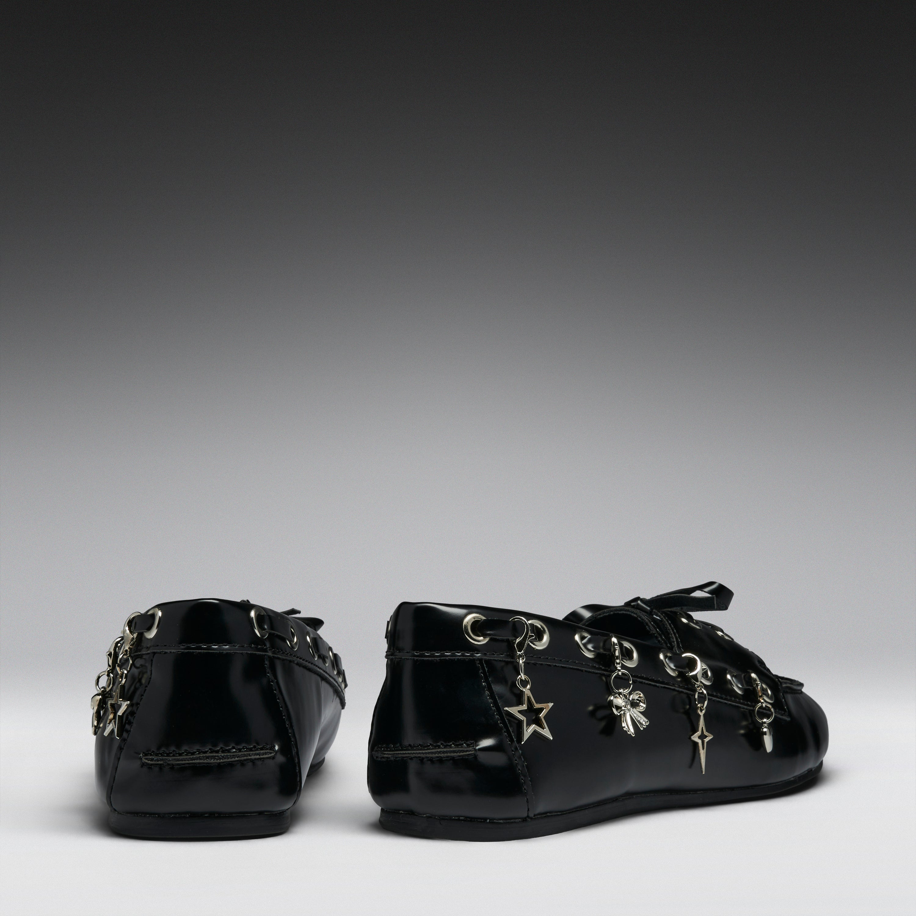 A pair of black patent leather lace-up flats decorated with silver-tone metal charms, including a star, cross, and clover, shown from the back and side against a dark gray gradient background.