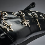 Close-up of a glossy black patent leather bag featuring silver-tone dangling charms shaped like a star, a bow, and a lightning bolt, with matching silver eyelets and studs.
