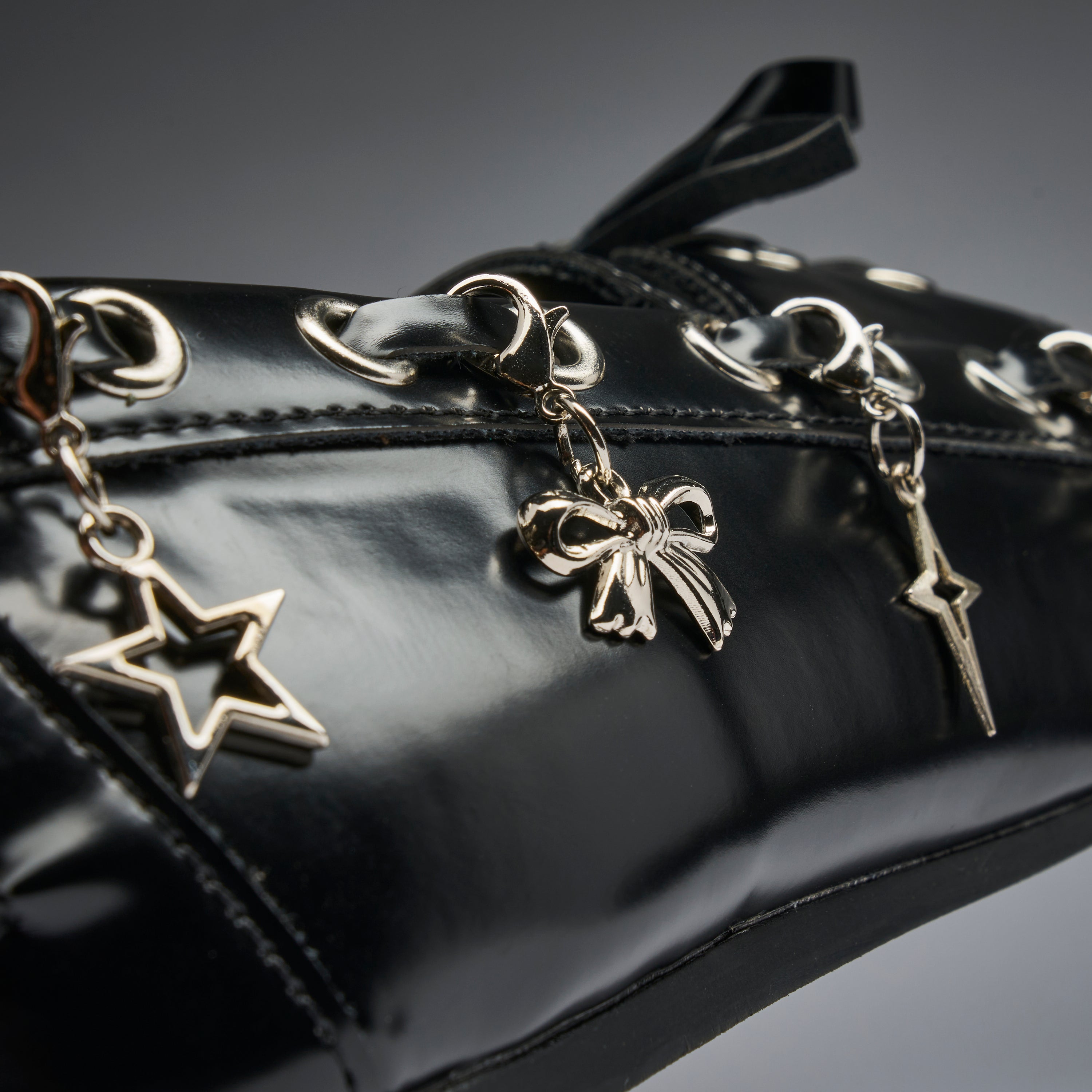 Close-up of a glossy black patent leather bag featuring silver-tone dangling charms shaped like a star, a bow, and a lightning bolt, with matching silver eyelets and studs.