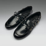 A pair of black leather boat-style loafers with decorative gold-tone hardware, bow ties, and dangling charms, displayed on a neutral gray gradient background. The brand name 'IOX' is visible on the insole.