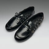 A pair of black leather boat-style loafers with decorative gold-tone hardware, bow ties, and dangling charms, displayed on a neutral gray gradient background. The brand name 'IOX' is visible on the insole.