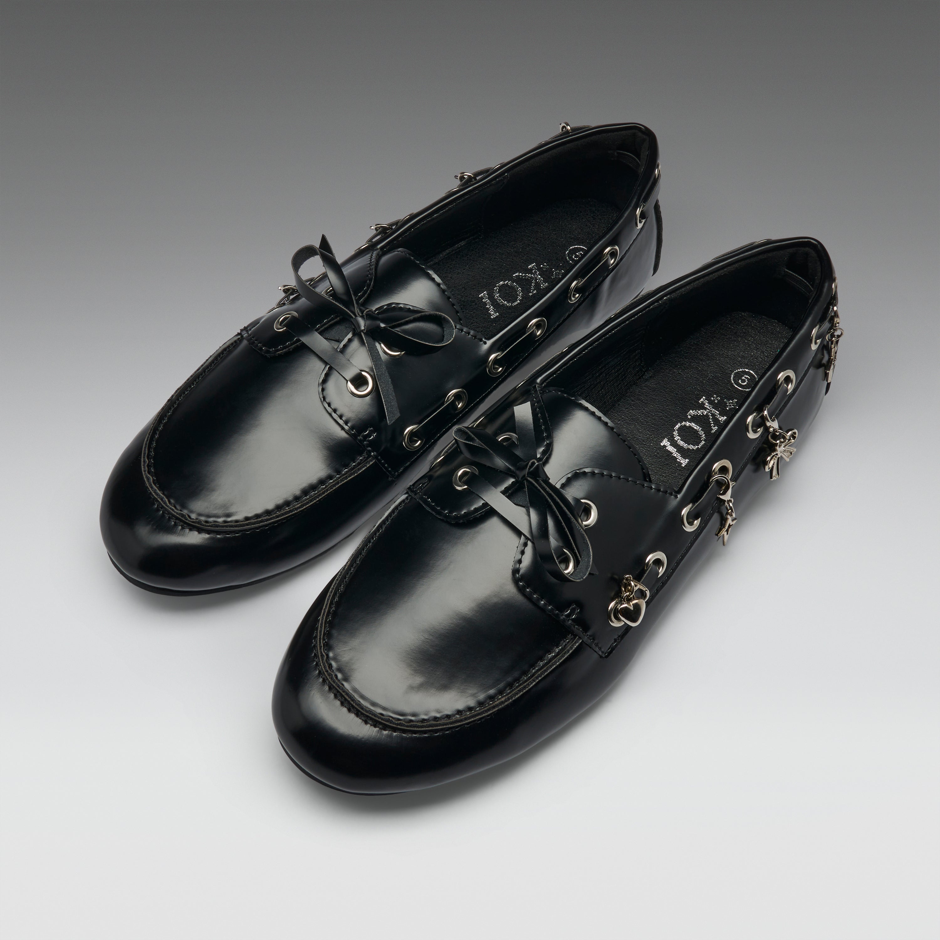 A pair of black leather boat-style loafers with decorative gold-tone hardware, bow ties, and dangling charms, displayed on a neutral gray gradient background. The brand name 'IOX' is visible on the insole.