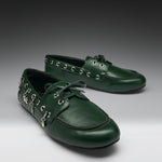 A pair of dark green leather boat-style shoes with decorative silver-tone metal eyelets, hanging charms, and matching green laces, displayed on a dark-to-light gray gradient studio background.