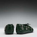 A pair of dark green leather baby booties decorated with silver-tone metal charms, including a star and bows, shown from the back on a gray gradient background.