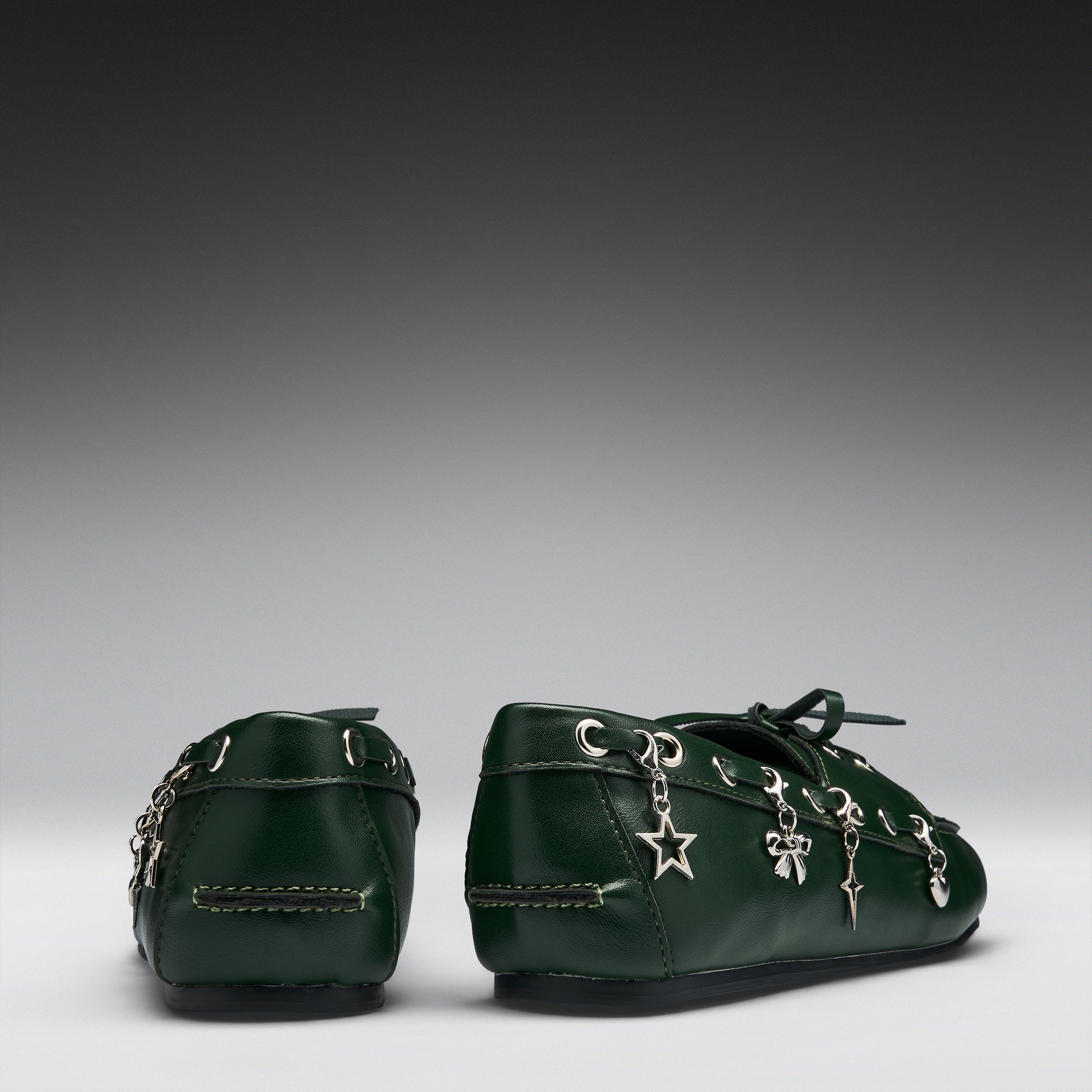 A pair of dark green leather baby booties decorated with silver-tone metal charms, including a star and bows, shown from the back on a gray gradient background.