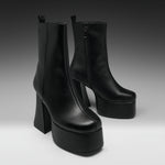 A pair of black leather ankle boots with high block heels and thick platform soles, photographed on a dark-to-light gray studio background.