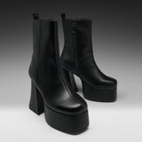 A pair of black leather ankle boots with high block heels and thick platform soles, photographed on a dark-to-light gray studio background.
