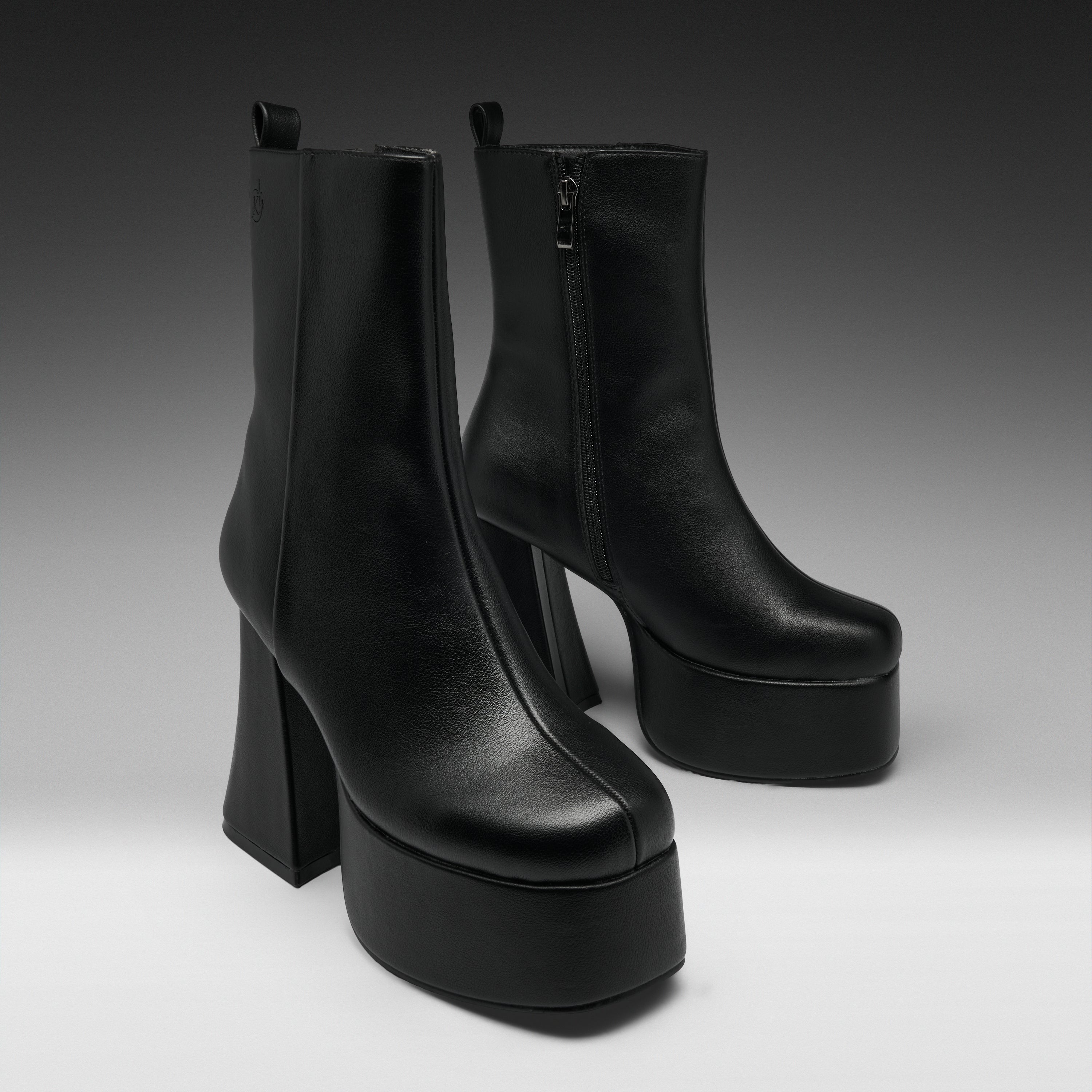 A pair of black leather ankle boots with high block heels and thick platform soles, photographed on a dark-to-light gray studio background.