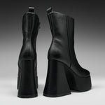 A pair of black leather ankle boots with chunky platform heels, shown from the back and side against a dark gray gradient studio background.
