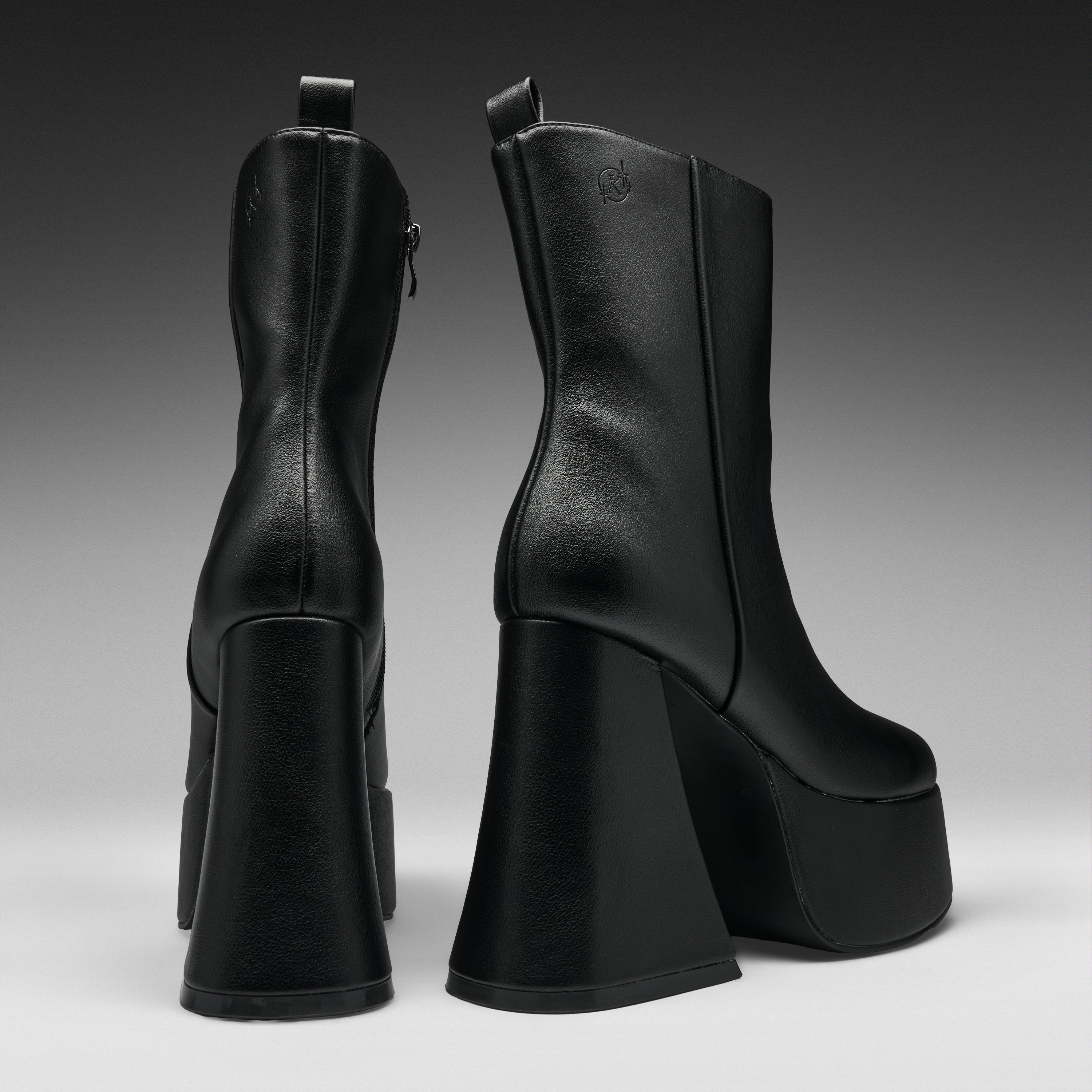 A pair of black leather ankle boots with chunky platform heels, shown from the back and side against a dark gray gradient studio background.