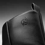 Close-up of a black textured leather bag or pouch with an embossed circular logo featuring a stylized letter K, set against a dark gray gradient background.