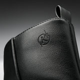 Close-up of a black textured leather bag or pouch with an embossed circular logo featuring a stylized letter K, set against a dark gray gradient background.