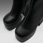 Close-up of a pair of black leather platform ankle boots with a chunky block heel, showing the toe and side profile against a neutral gray background.