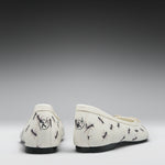 A pair of off-white ballet flats with a black ant print pattern, shown from the back against a dark-to-light gray gradient background.
