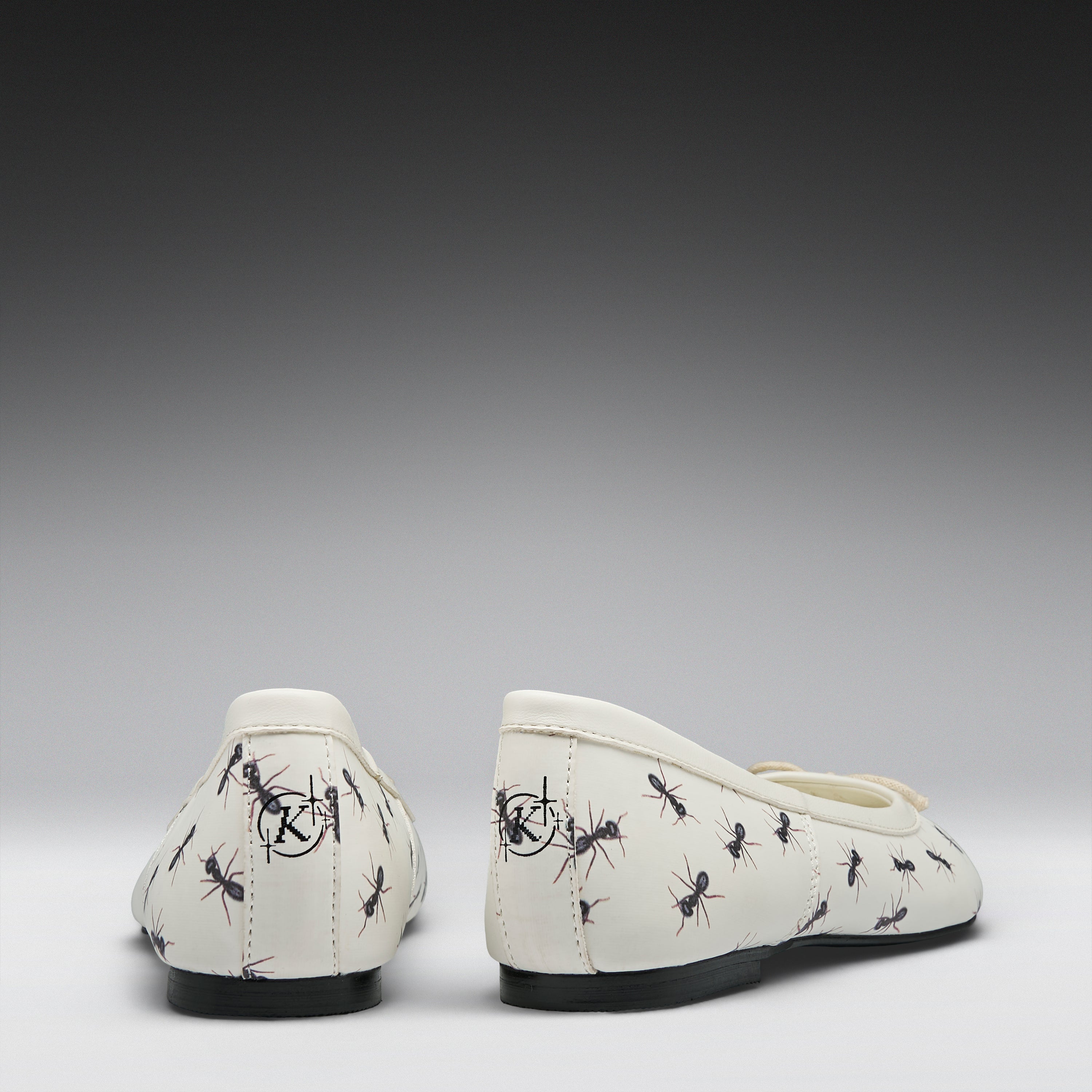 A pair of off-white ballet flats with a black ant print pattern, shown from the back against a dark-to-light gray gradient background.
