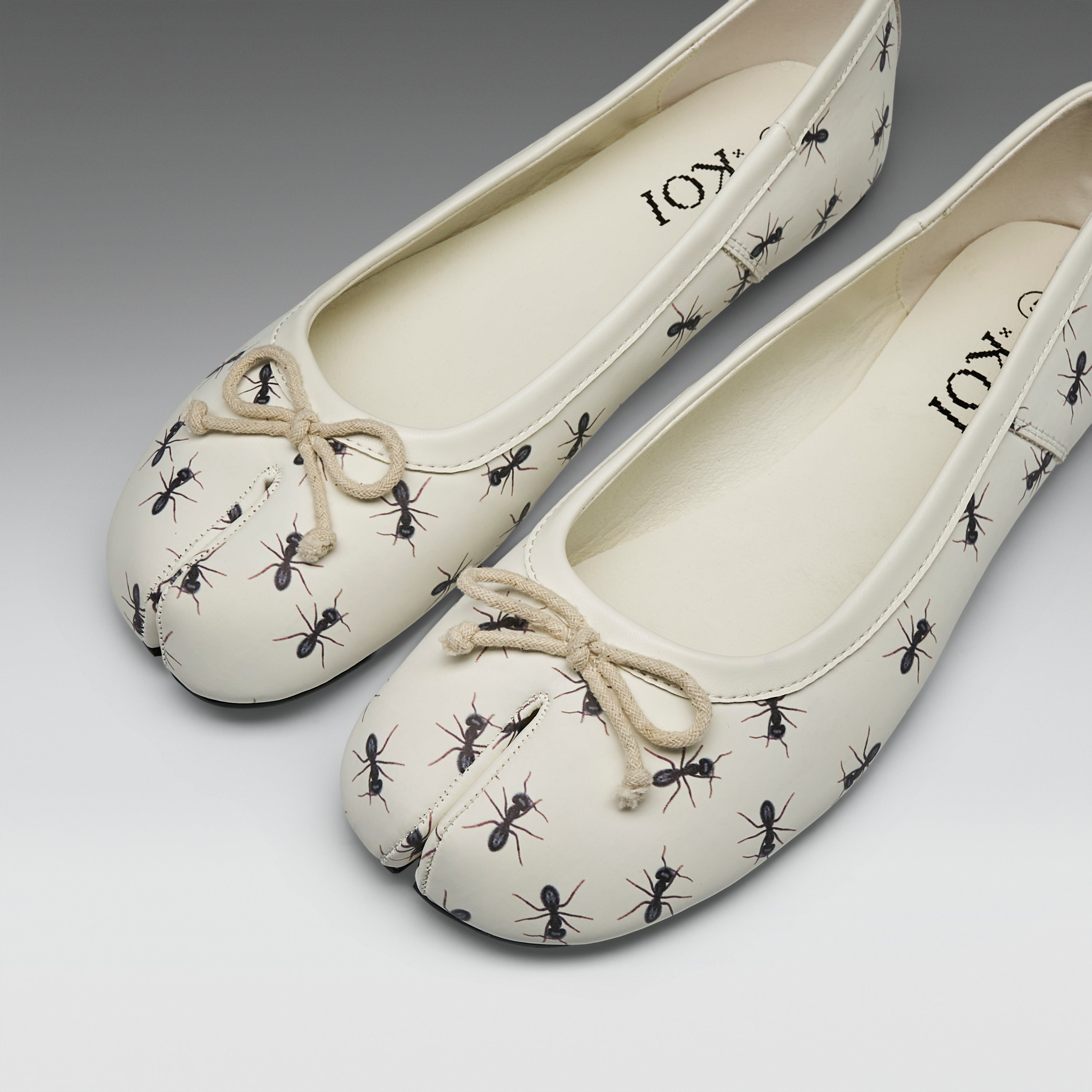 A pair of cream-colored split-toe ballet flats with a black ant print and a beige rope bow on the toe, shown on a neutral gray background.