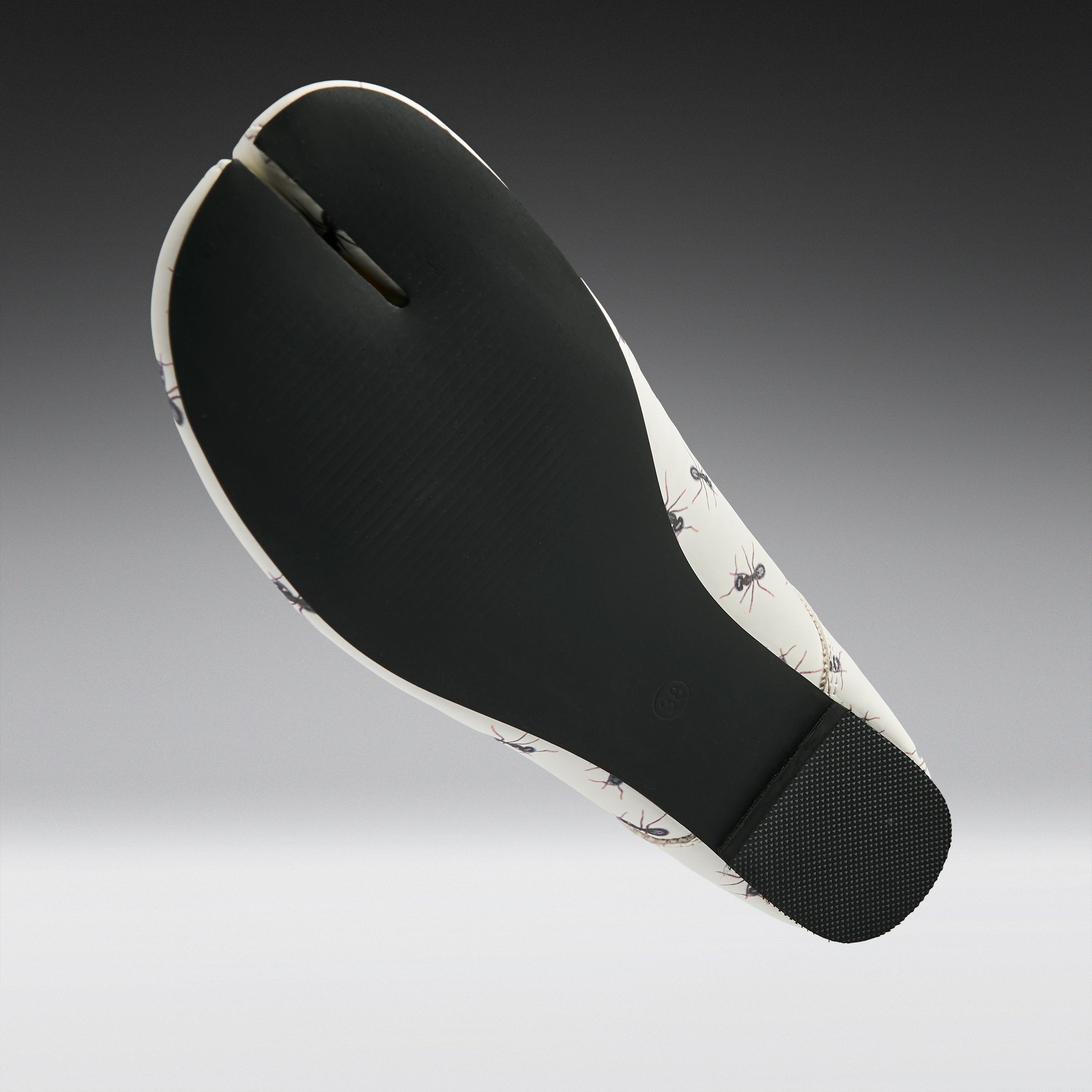 A split-foot flat sandal shown from the sole view, featuring a black textured midfoot and heel with a white and black ant-patterned outsole, set against a dark-to-light gray gradient background.