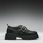 A black leather platform shoe with double buckle straps, contrast white stitching, and a chunky lug sole, shown in a side profile on a gray gradient background.