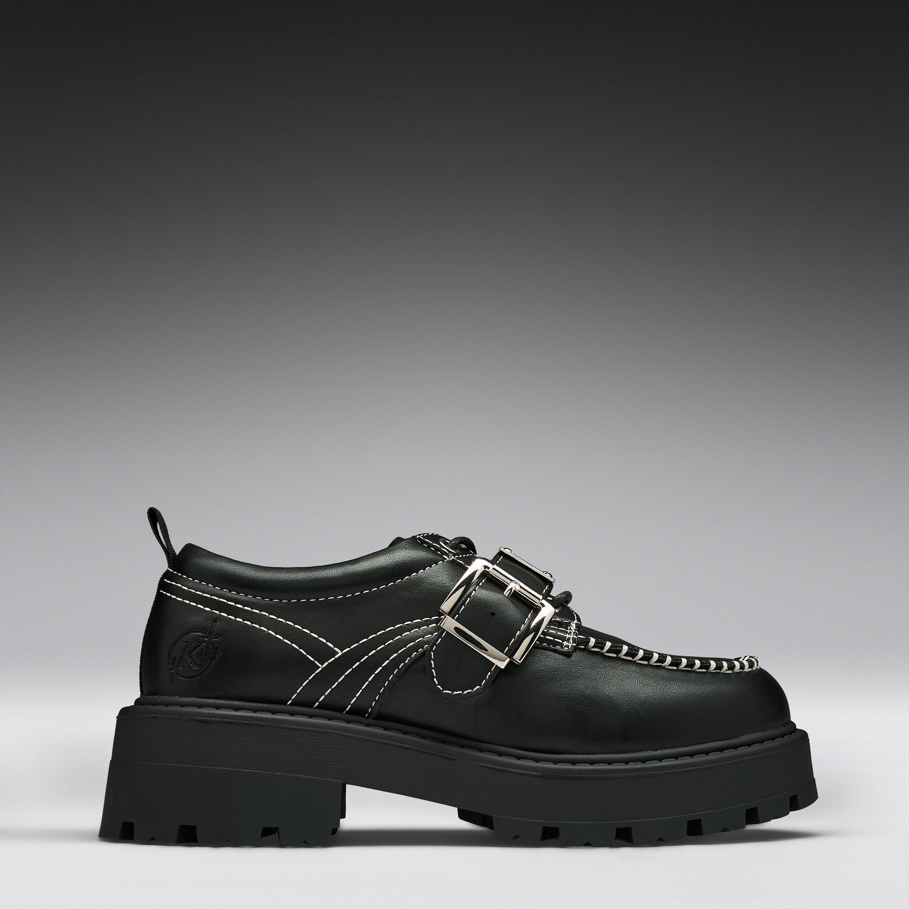 A black leather platform shoe with double buckle straps, contrast white stitching, and a chunky lug sole, shown in a side profile on a gray gradient background.